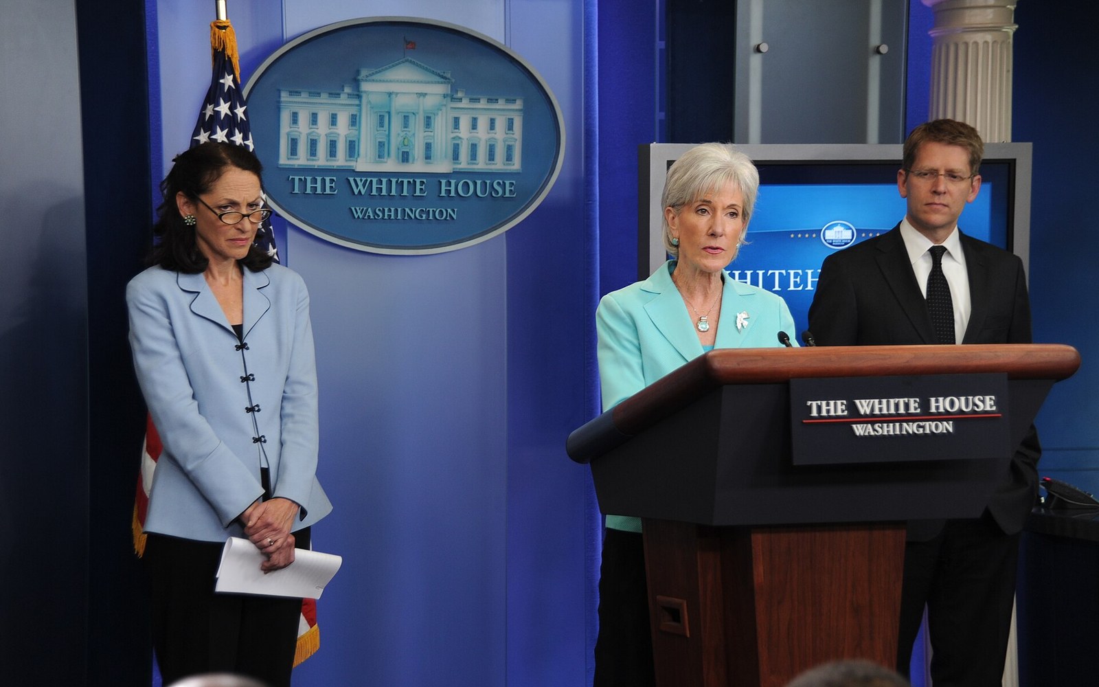 Two women and a man stand at a White House press briefing room podium